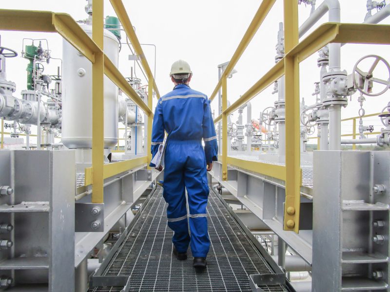 Radnik u plavom zaštitnom odelu i šlemu hoda metalnim prolazom u industrijskom postrojenju okružen cevima i ventilima.